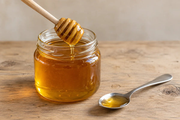 Cuchara de madera con ranuras y cucharilla de acero apoyadas junto a un tarro de miel