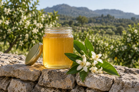 Tarro de miel de azahar dorada junto a flores de naranjo, sobre un muro de piedra con bancales mediterráneos desenfocados